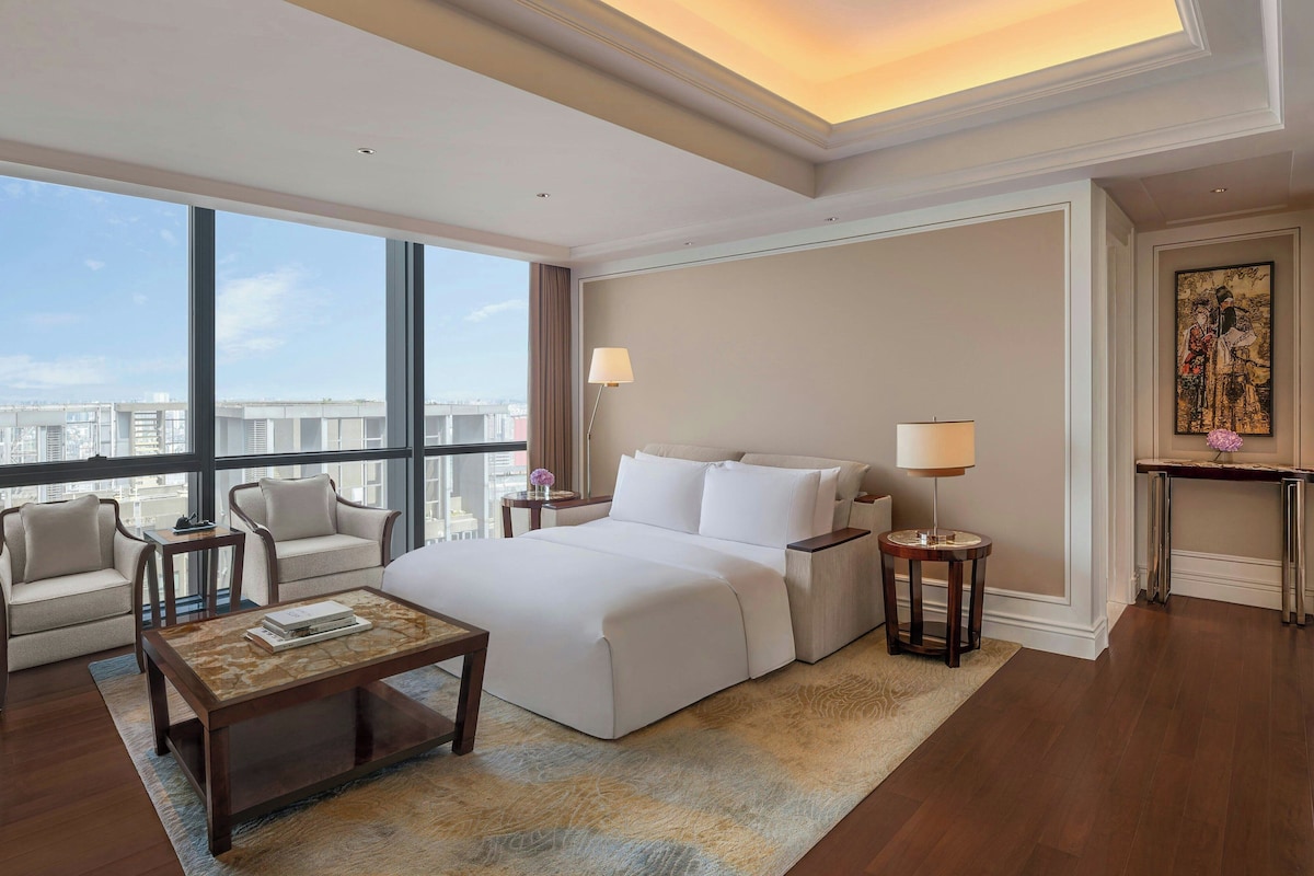 A hotel room with a large bed, a sitting area with a chair and a small table, a nightstand with a lamp, and a view of the city through large windows.