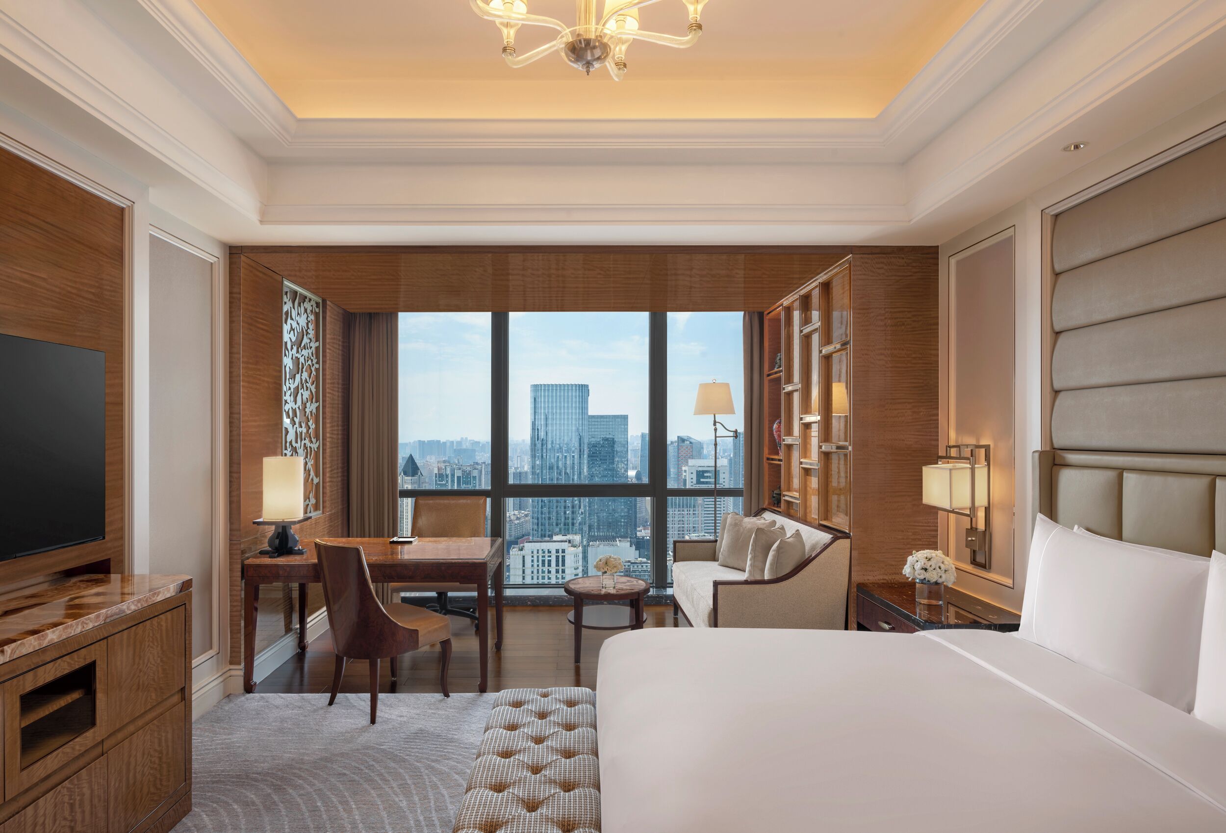 A hotel room with a large bed, a desk, a chair, a TV, and a view of the cityscape.