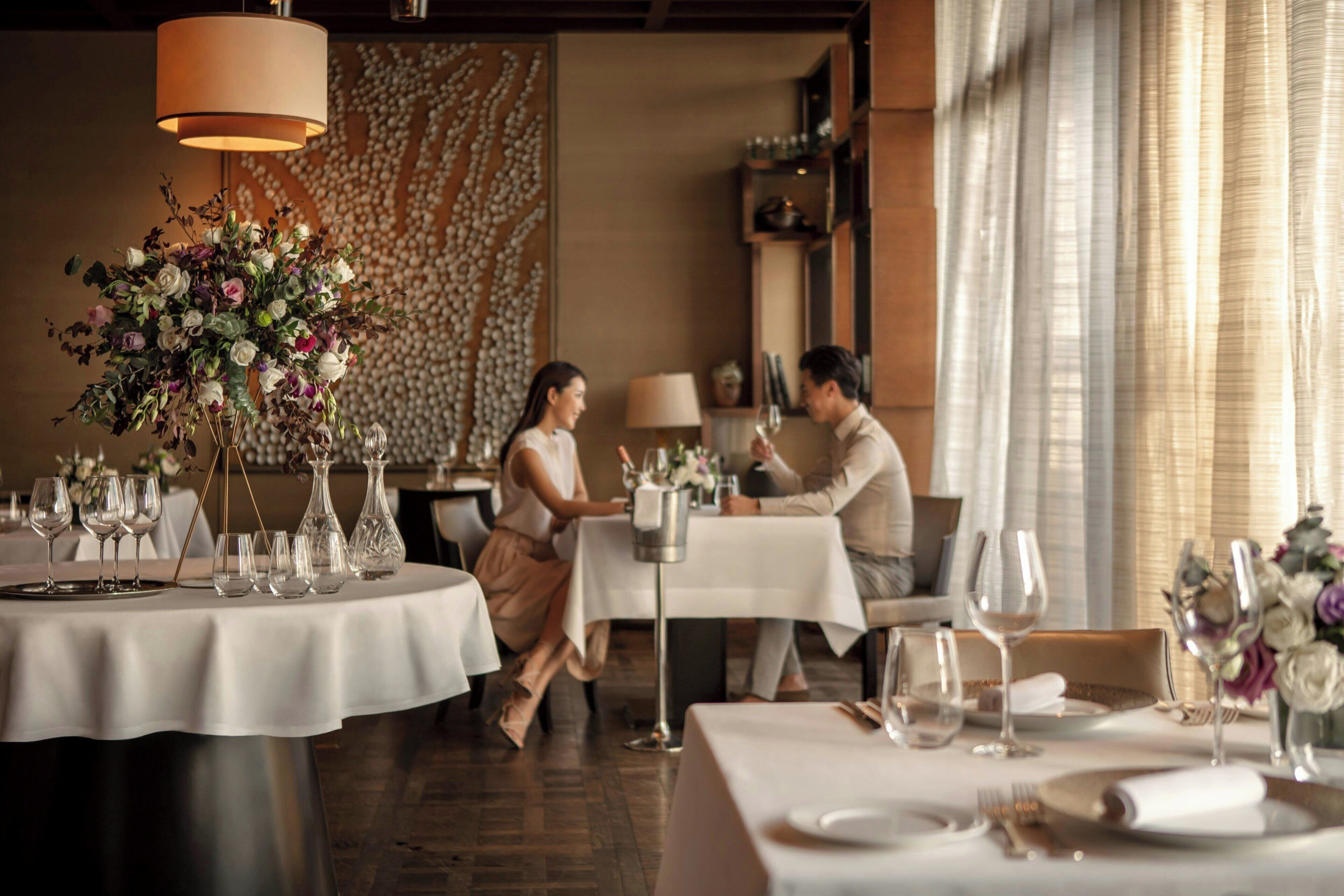 A restaurant interior with round tables set for dining, a floral centerpiece, and a wall with a textured pattern.
