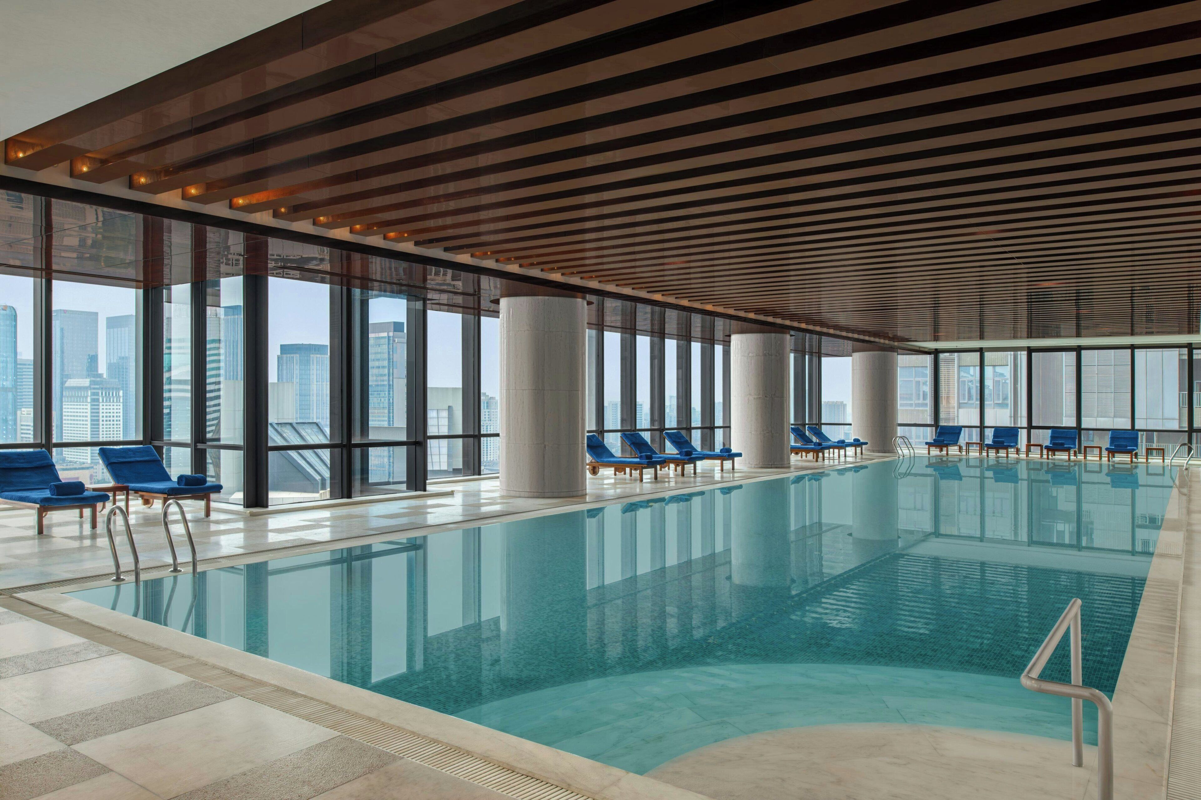 Una piscina interior con paredes de vidrio transparente, sillones reclinables azules y vistas a la ciudad.