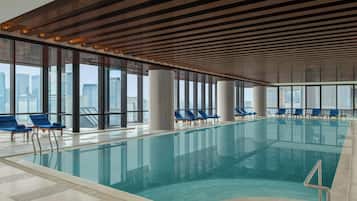 Une piscine intérieure avec des parois en verre transparent, des fauteuils bleus et une vue sur la ville.