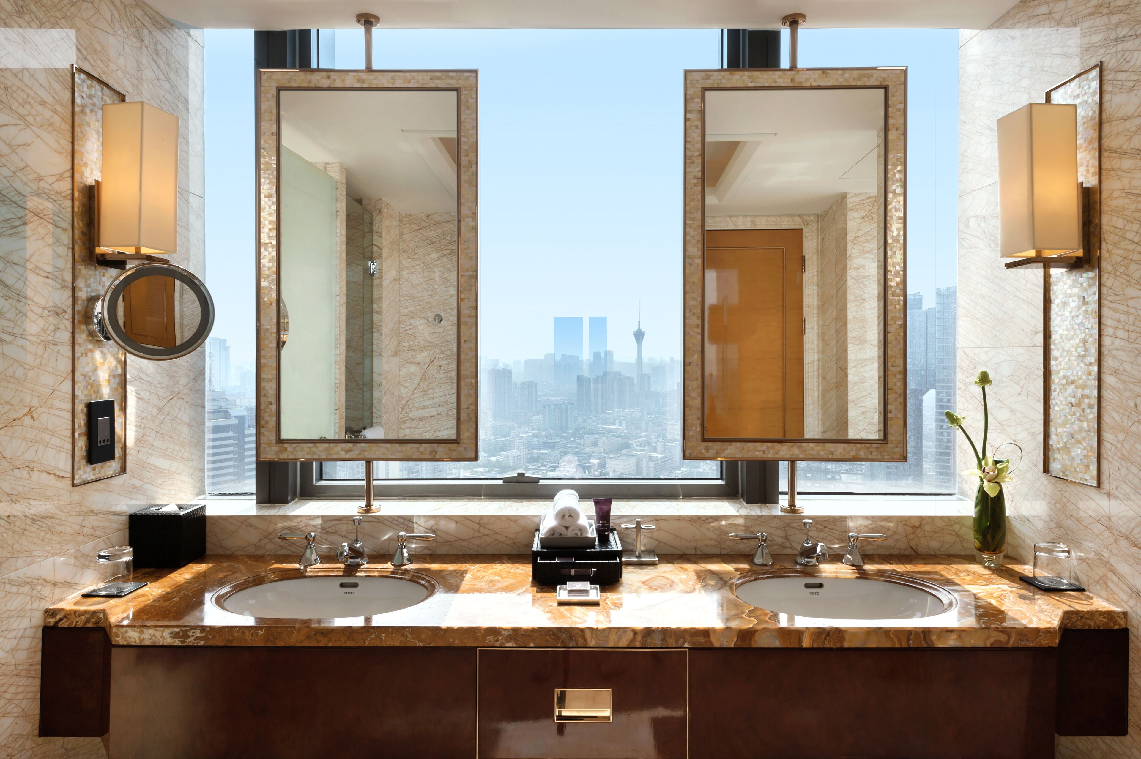 A modern bathroom with two sinks, large mirrors, and a view of a cityscape through a large window.