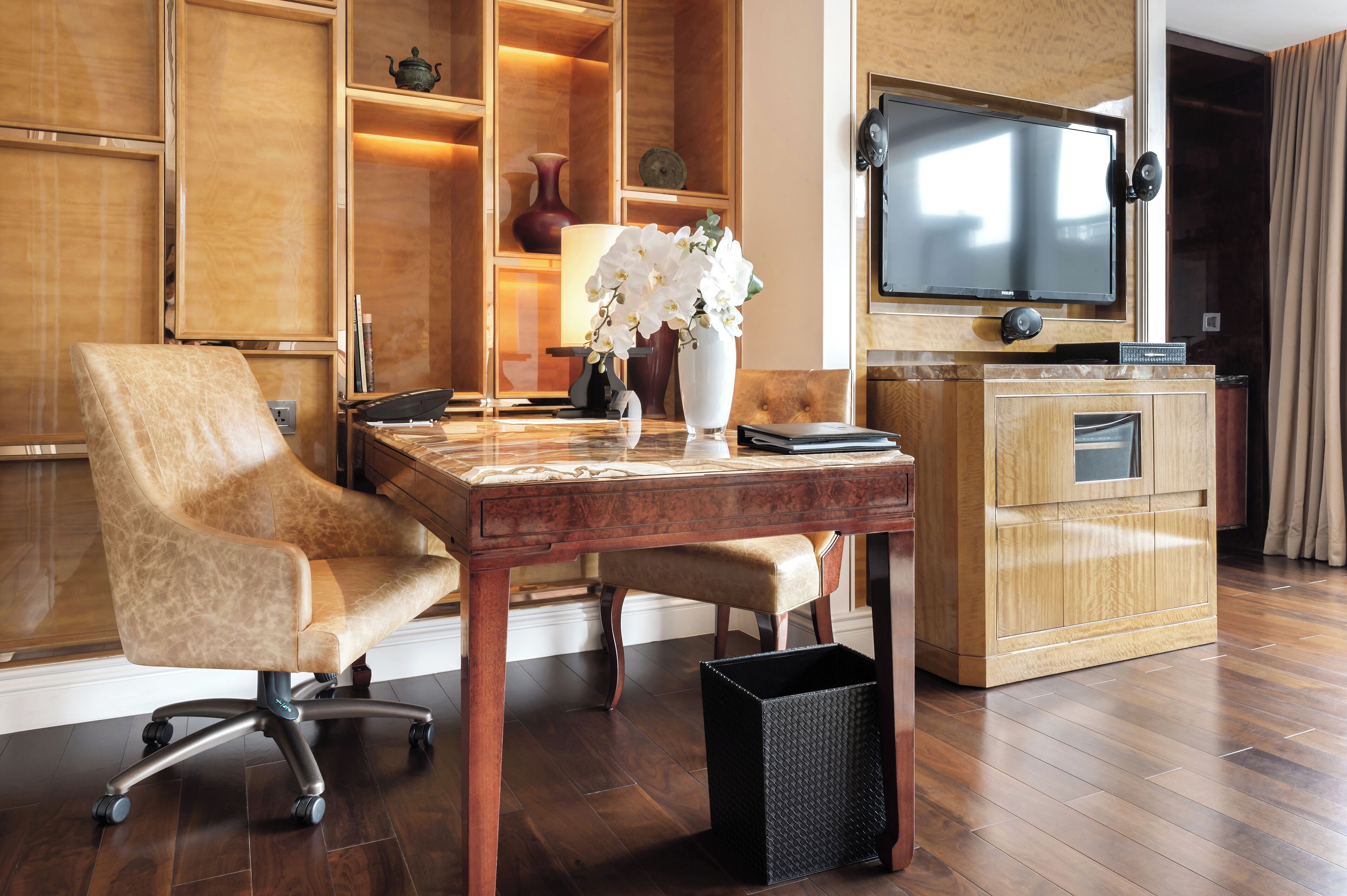 Una habitación de hotel moderna con un escritorio de madera, un sillón cómodo, un televisor montado en la pared y un jarrón con flores blancas.