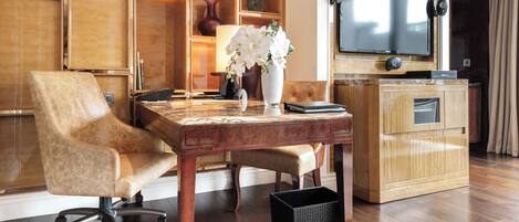 Una habitación de hotel moderna con un escritorio de madera, un sillón cómodo, un televisor montado en la pared y un jarrón con flores blancas.