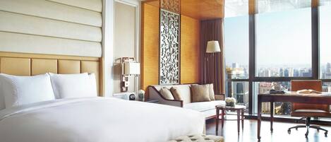 Habitación de hotel con una cama grande, un sofá, un escritorio con una silla y vistas a la ciudad a través de amplios ventanales.