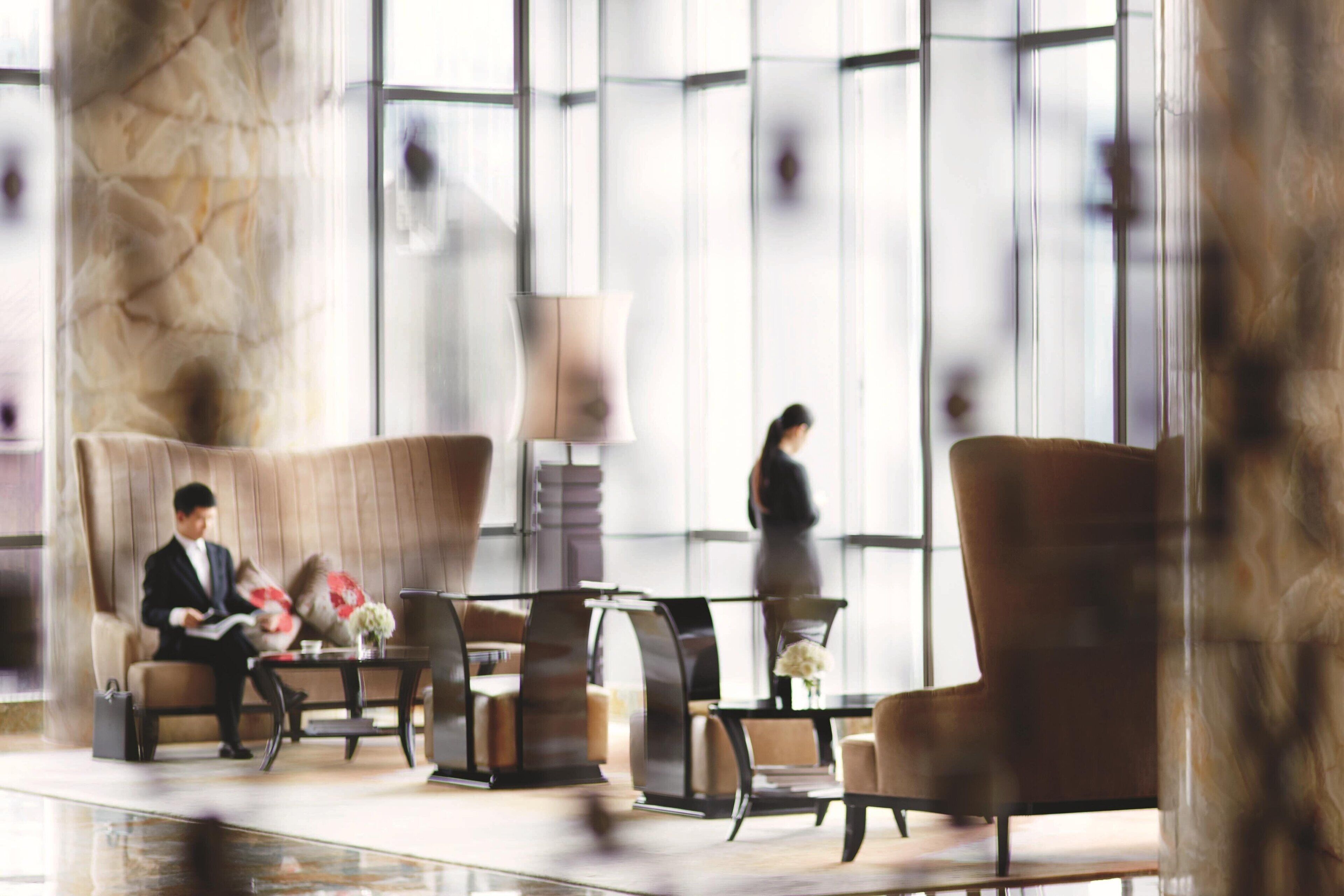 A modern lobby with a man reading a book, a woman walking, and a seating area with a lamp and flowers.
