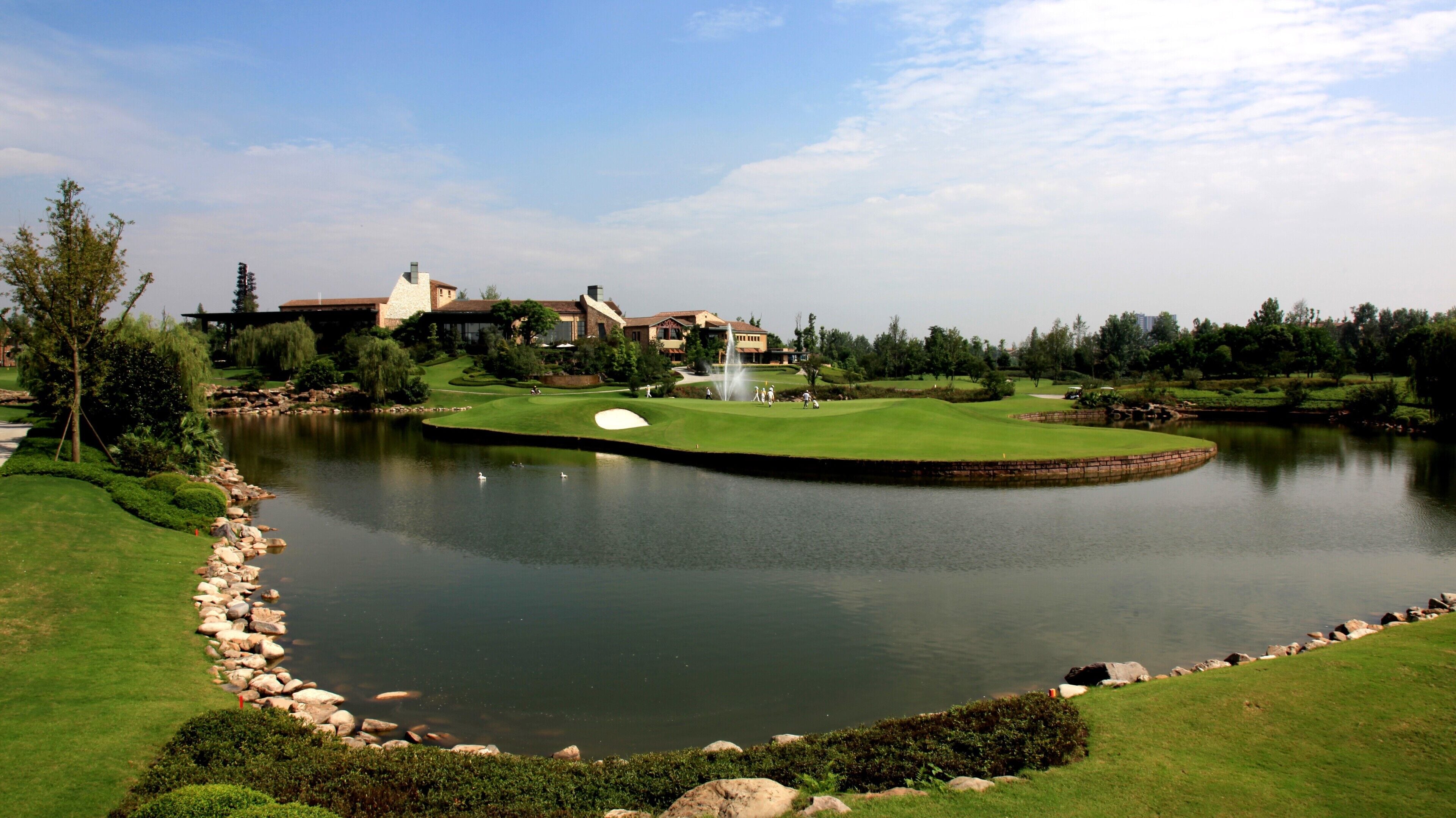 A golf course with a pond, a fountain, and a clubhouse in the background.