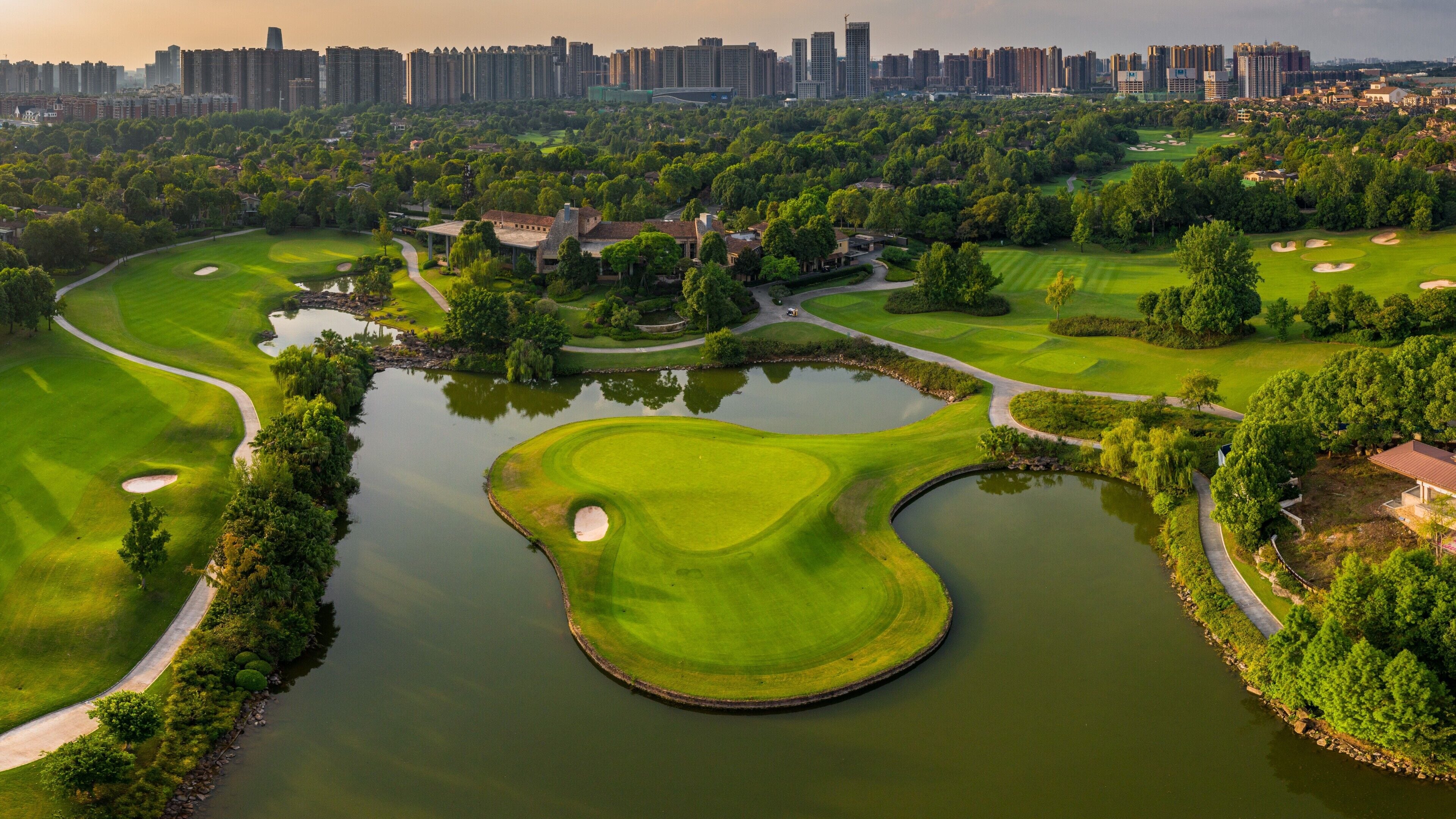 A golf course with a pond, surrounded by trees and buildings in the background.