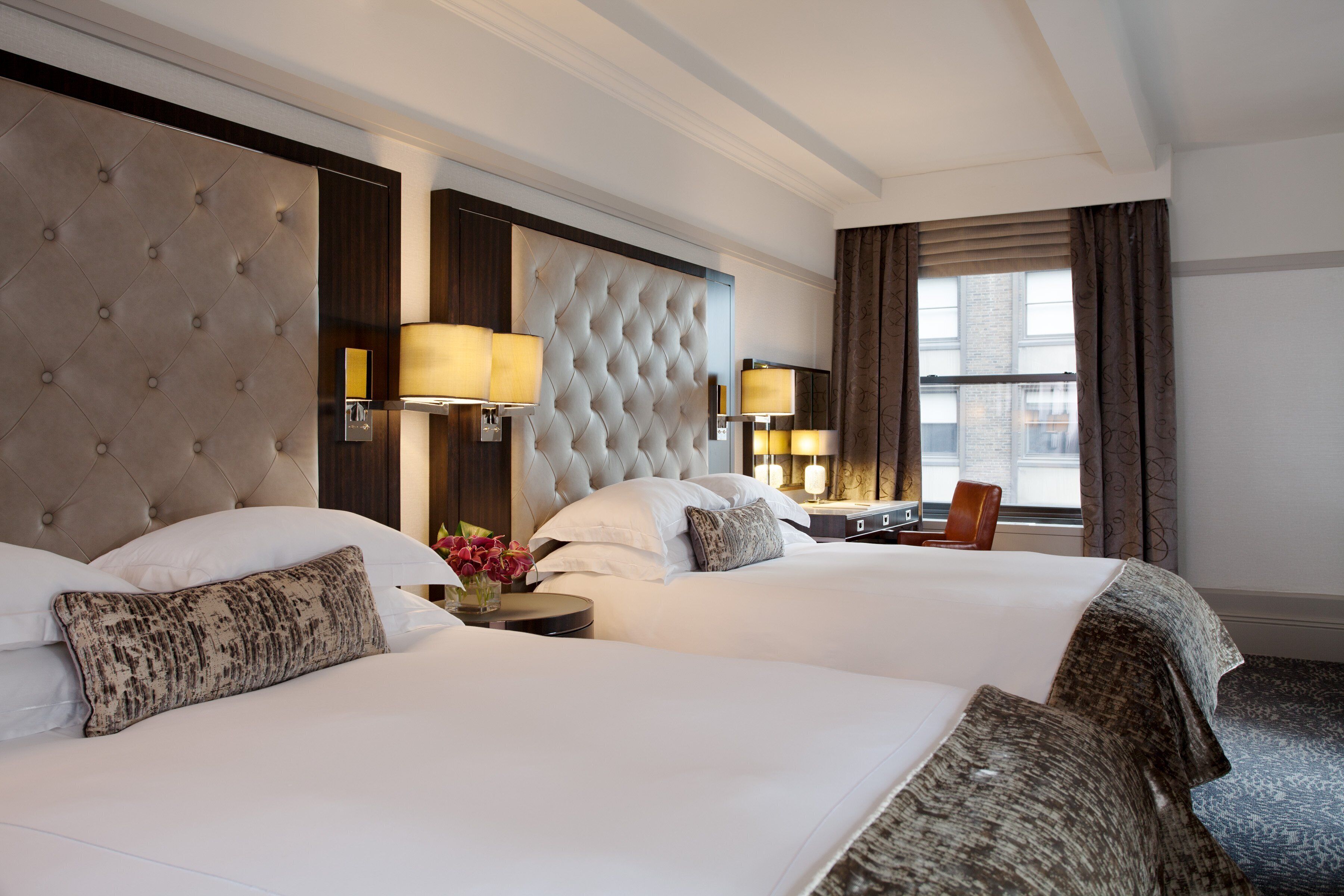 A hotel room with two large beds, a tufted headboard, bedside lamps, a small table with a vase of flowers, and a window with curtains.