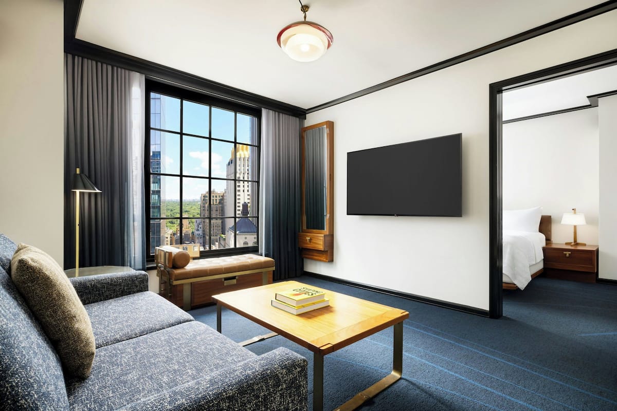 Ein modernes Hotelzimmer mit großem Fenster, Flachbildfernseher, einer Couch, einem Couchtisch und einem Nachttisch mit Lampe.