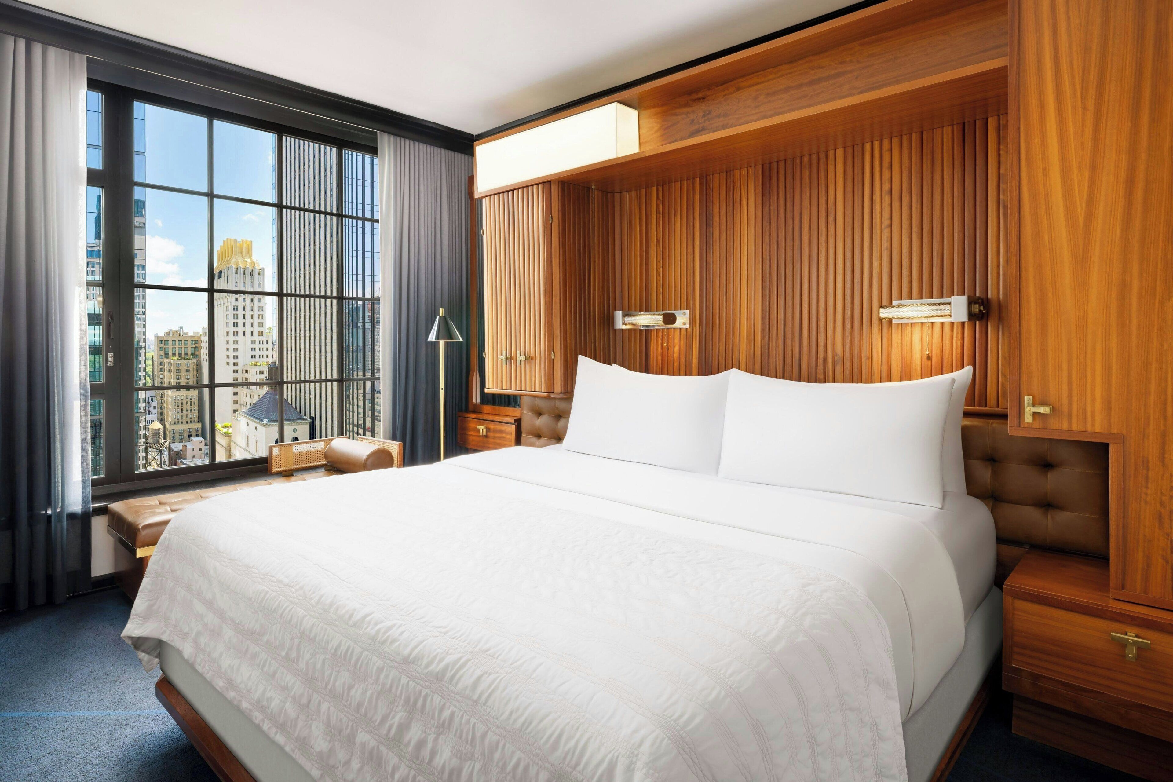 A hotel room with a large bed, wooden paneling, a view of city buildings, a nightstand, and a lamp.