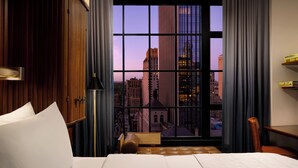 A hotel room with a large window offering a city view, a bed with white linens, a wooden headboard, and a small seating area with a bench and a chair. - Le Meridien New York, Central Park by Marriott (New York)
