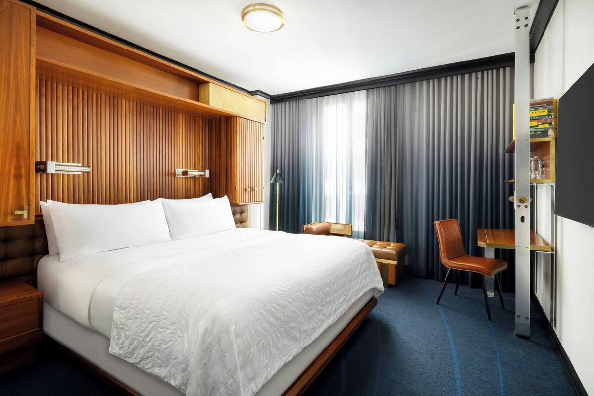 Ein Hotelzimmer mit einem großen Bett, einem Holzkopfteil, einem Schreibtisch mit Stuhl, einem Bücherregal und einem großen Fenster mit Vorhängen.