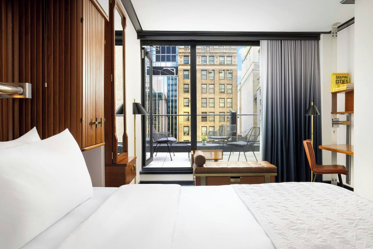 Ein Hotelzimmer mit einem großen Bett, einem Schreibtisch, einem Stuhl und Blick auf die Stadt durch ein großes Fenster.