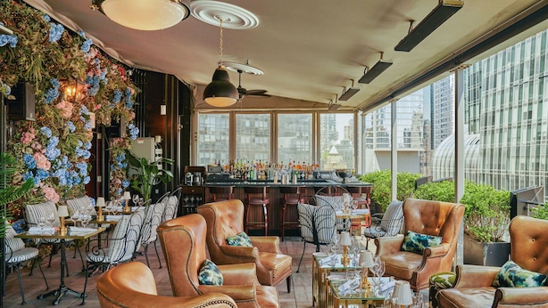 A bar area with leather chairs and tables, a large window with a city view, and a wall decorated with flowers. - Le Meridien New York, Central Park by Marriott (New York)