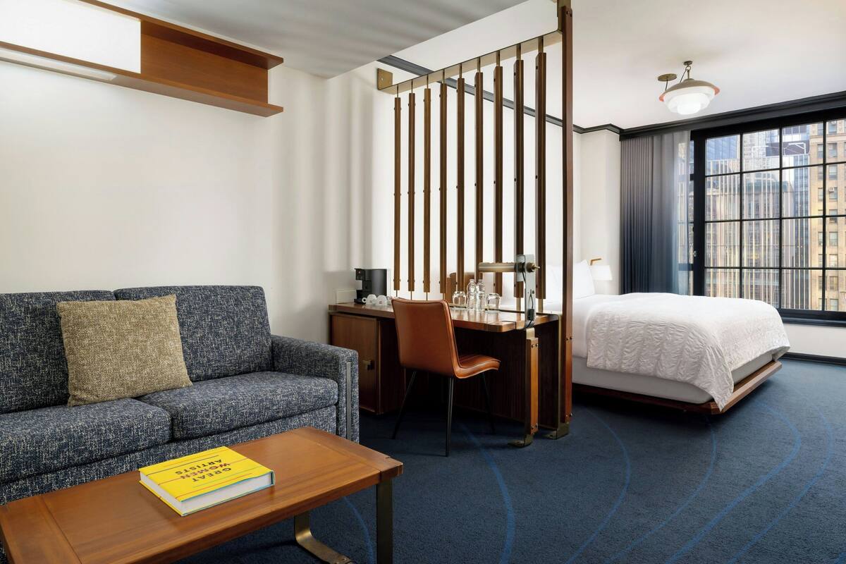 Ein Hotelzimmer mit Sofa, Schreibtisch, Bett und Blick auf die Stadt durch große Fenster.