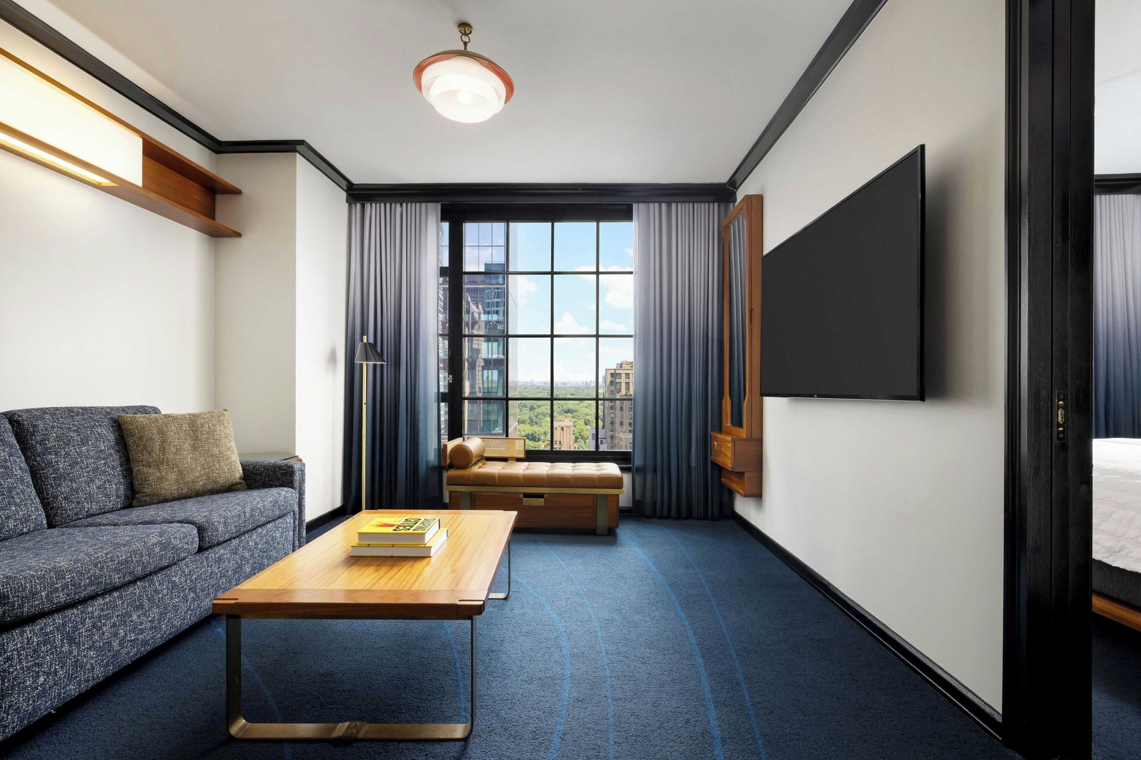 Une chambre d’hôtel moderne dotée d’un canapé, d’une table basse en bois, d’un téléviseur à écran plat et d’une grande fenêtre avec des rideaux.