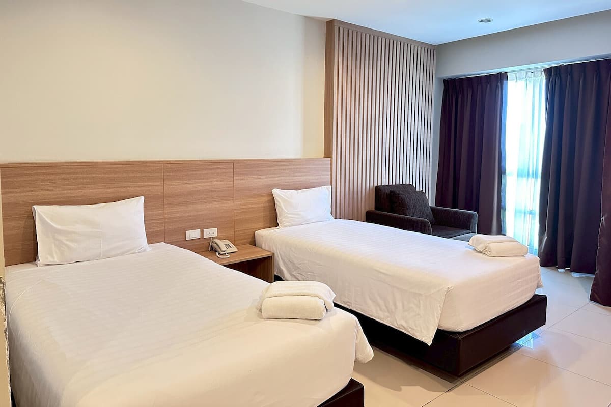 Habitación de hotel con dos camas, una mesita de noche con teléfono, una silla y un ventanal con cortinas.