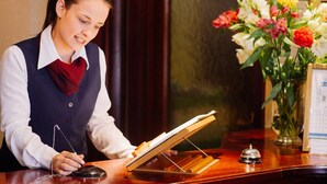 A reception desk with a person holding a clipboard, a telephone, and a bell. There is a vase with flowers and a framed menu on the desk. - Stafford Hotel (St. Helier)