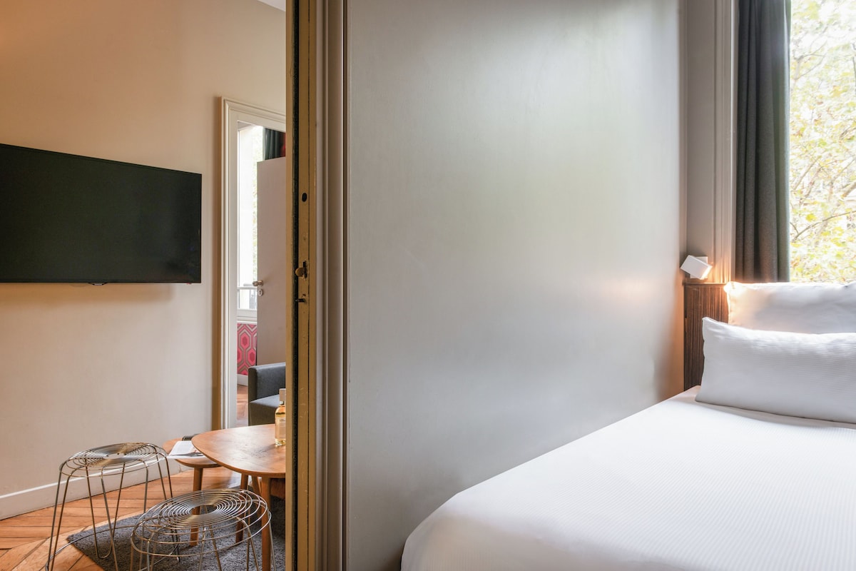 A hotel room with a bed, a TV mounted on the wall, a dining area with chairs, and a view of the outdoors.