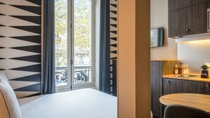 Habitación de hotel con cama, ventana con cortinas, vista al exterior, una kitchenette pequeña con lavamanos y un televisor montado encima.
