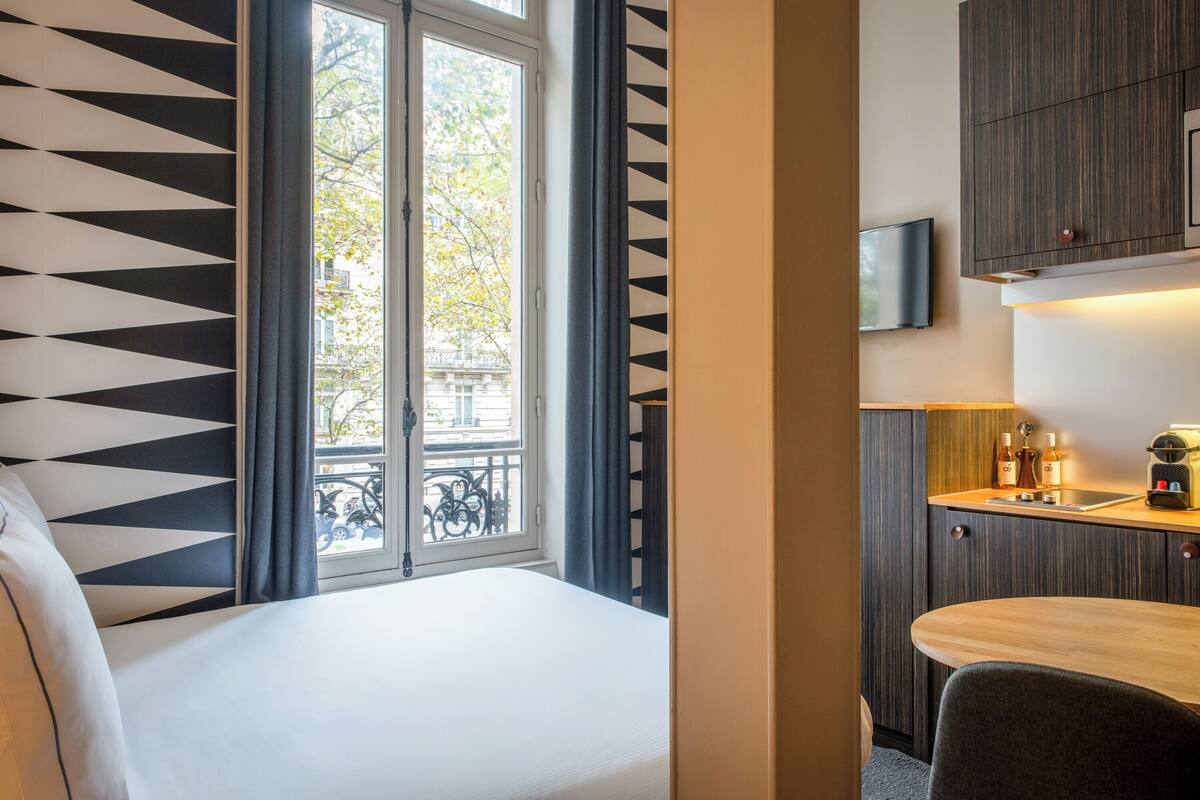 A hotel room with a bed, a window with curtains, a view of the outside, a small kitchenette with a sink and a television mounted above it.