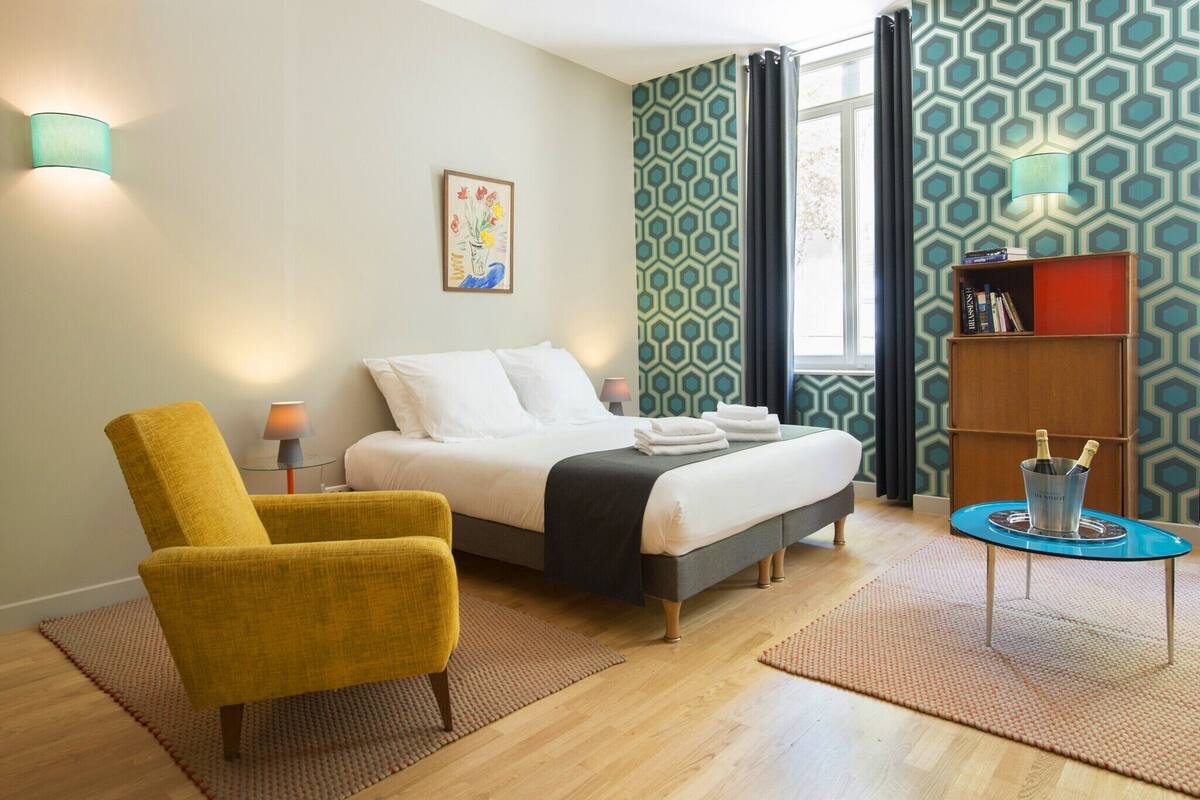 A hotel room with a bed, a yellow armchair, a small table with a lamp, a bookshelf, and patterned wallpaper.