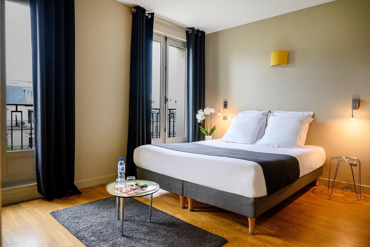 A hotel room with a bed, a small table with a water bottle, a potted plant, and a window with curtains.