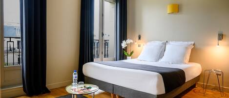Quarto de hotel com cama, uma pequena mesa com uma garrafa de água, uma planta em vaso e vista para o exterior.