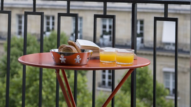 Un balcón con una mesa que sostiene croissants, un libro y vasos de jugo de naranja, con vistas a un edificio de múltiples ventanas y techo gris.