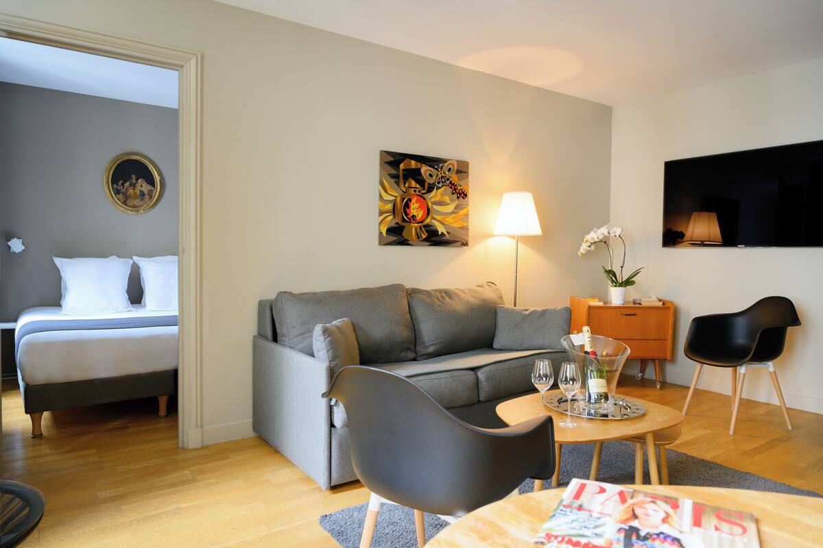 A modern living room with a grey sofa, a black armchair, a coffee table with a magazine, a lamp, and a painting on the wall.
