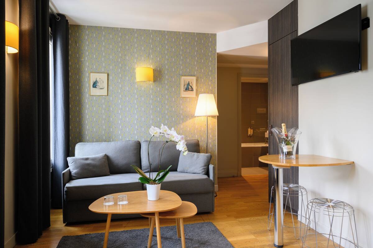 A modern living room with a grey sofa, two small round tables, a potted orchid, and a wall-mounted TV.