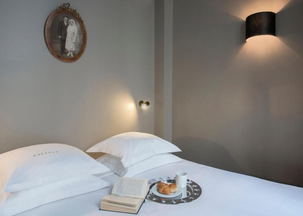 A neatly made bed with white linens, a framed photograph on the wall, a bedside table with a book, a cup, and a plate with pastries.