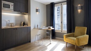 Una cocina moderna con microondas, hervidor y botella de vino, una pequeña mesa de comedor con sillas, y un sillón amarillo.