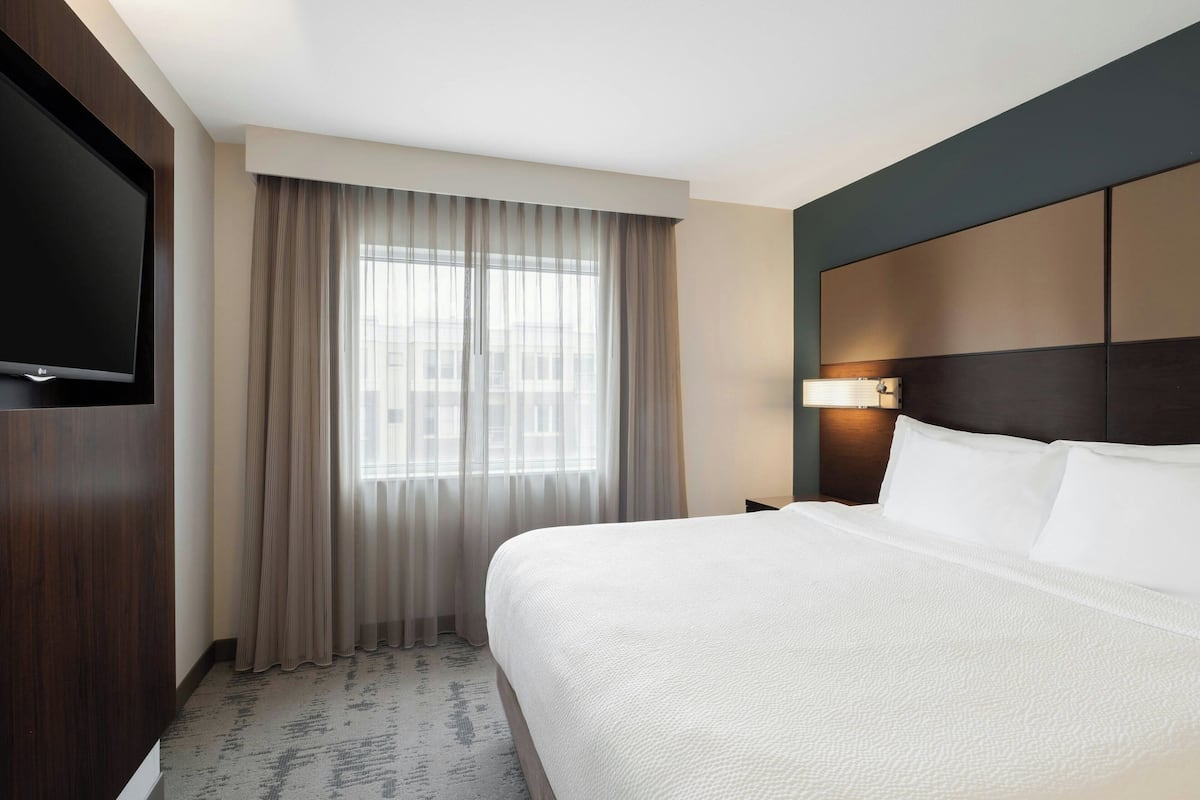 A hotel room with a large bed, a flat-screen TV mounted on the wall, a window with sheer curtains, and a nightstand with a lamp.
