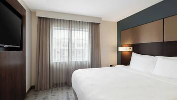Habitación de hotel con una cama grande, un televisor de pantalla plana montado en la pared, una ventana con cortinas translúcidas y una mesita de noche con una lámpara.