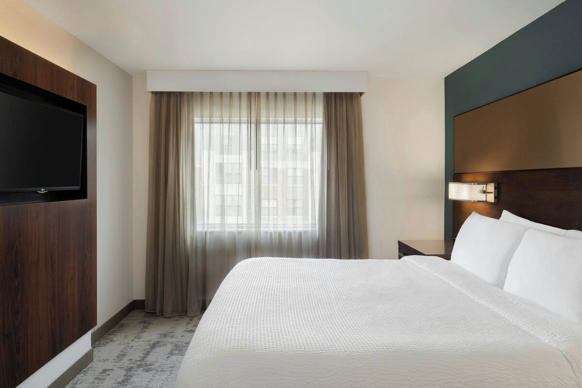 A hotel room with a large bed, a flat-screen TV mounted on the wall, a window with curtains, and a nightstand with a lamp.