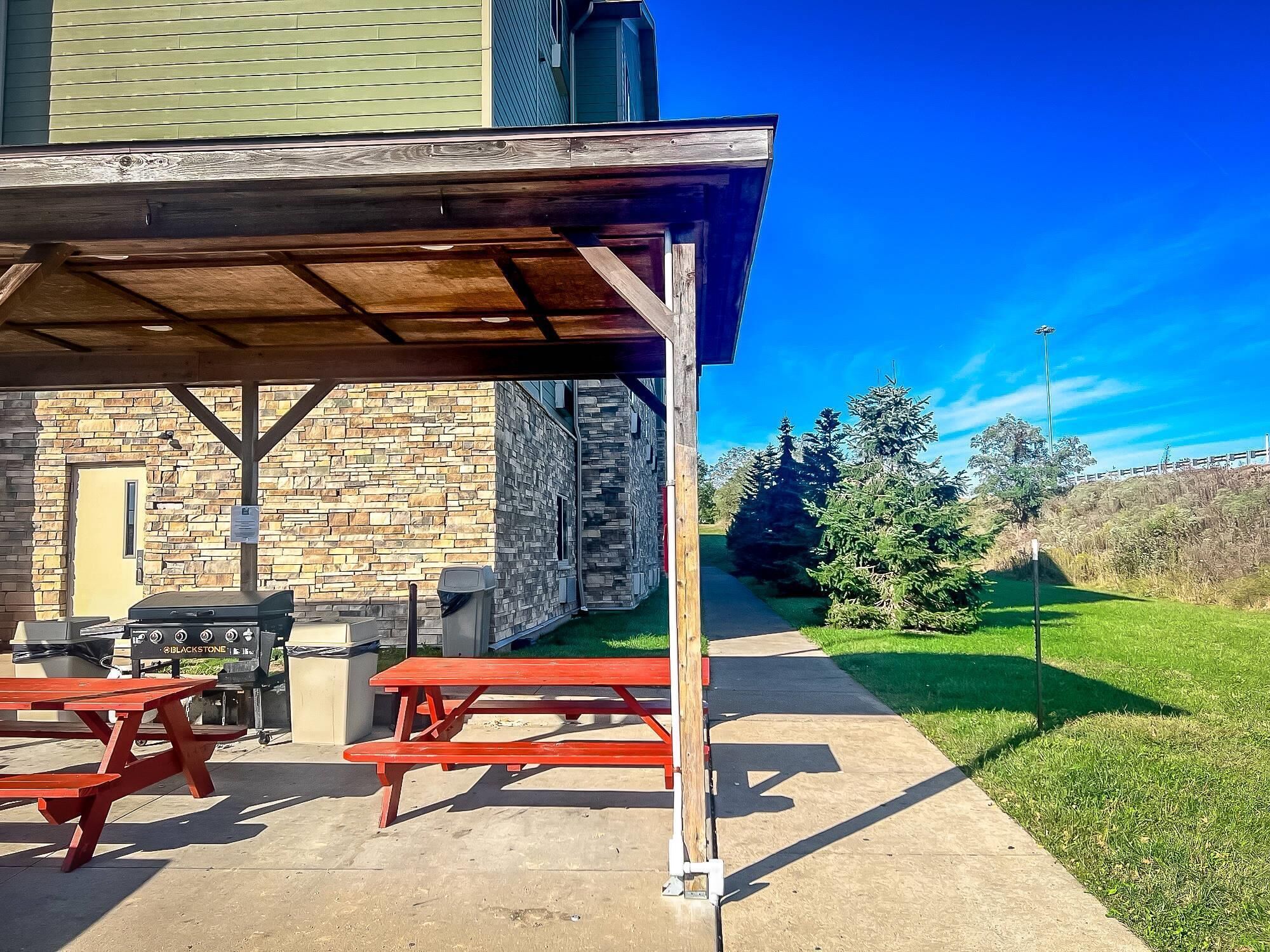 An outdoor patio with picnic tables, a barbecue grill, and a stone building in the background.