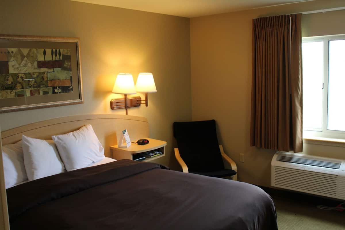 A hotel room with a bed, two bedside lamps, a chair, an air conditioner, and a window with curtains.