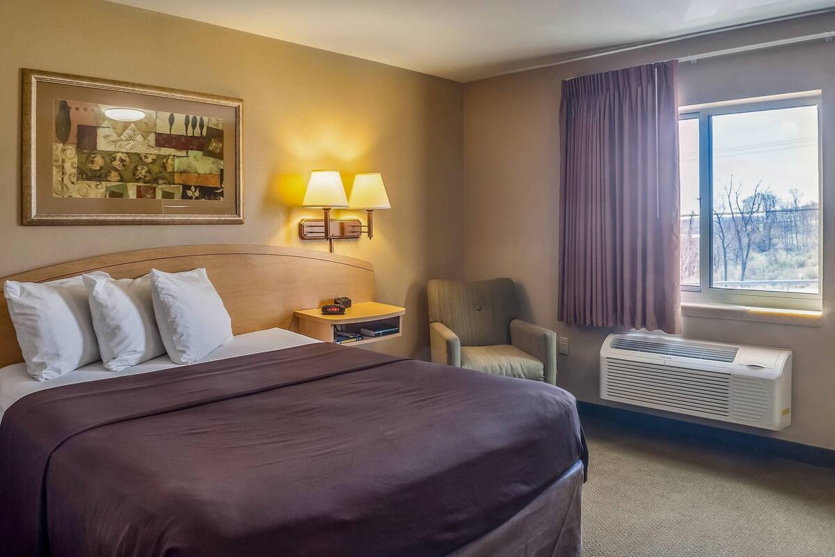 A hotel room with a bed, two wall-mounted lamps, a chair, a window with curtains, and an air conditioning unit.