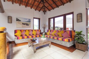 Una habitación con un banco de piedra, sofás coloridos a rayas, una mesa de centro de madera y grandes ventanas con vistas a la montaña.