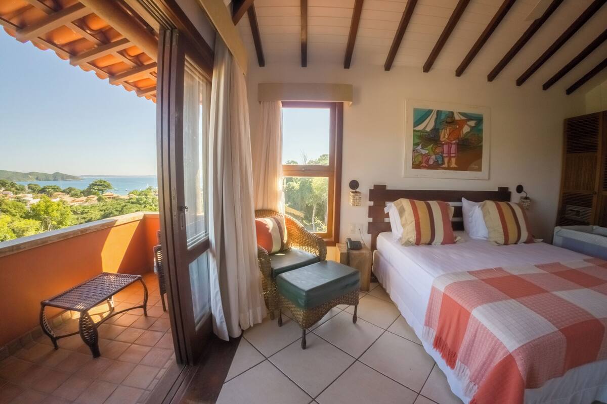 A bedroom with a bed, a small table, a bench, and a view of the sea.