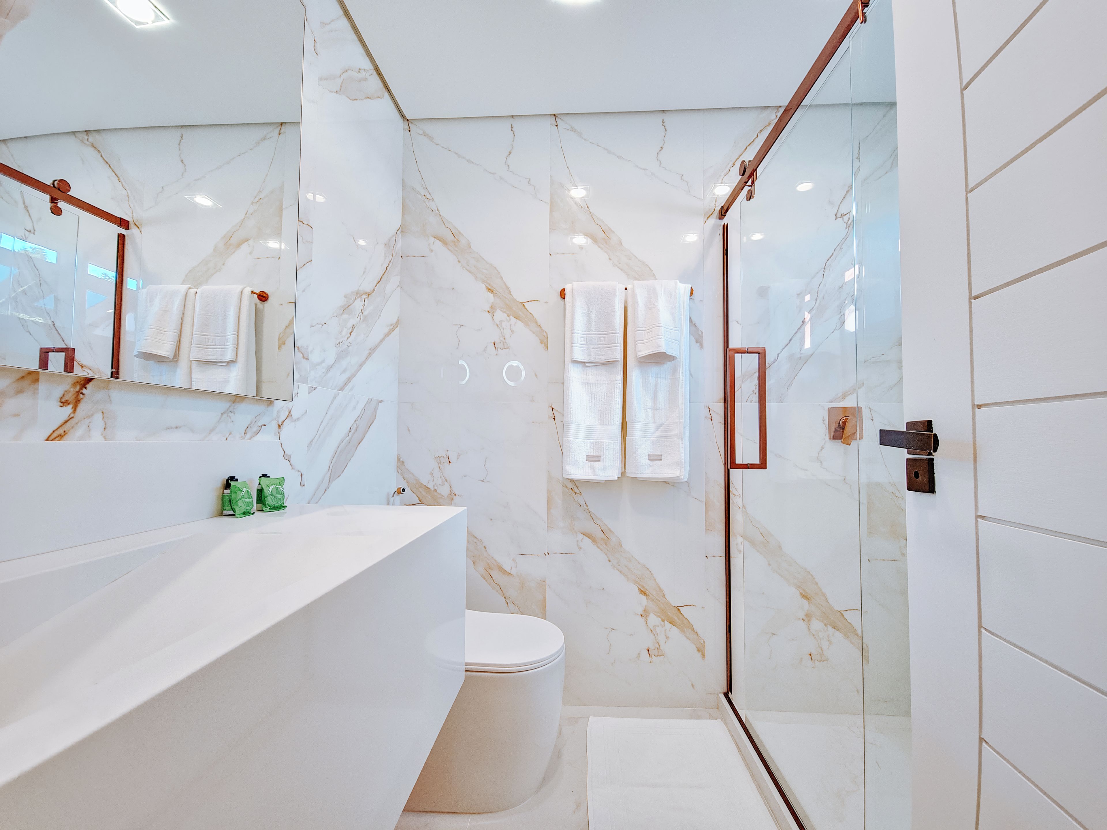 Un baño moderno con pared de mármol, cabina de ducha de cristal, inodoro blanco y tocador con espejo.