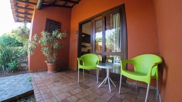 Un patio con dos sillas verdes y una mesa, una planta en maceta y una ventana con cortinas.