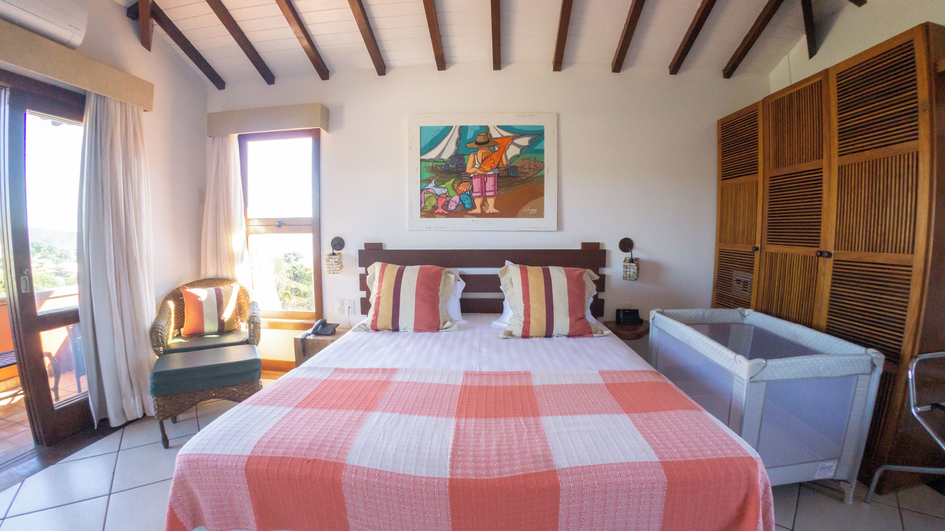 A hotel room with a bed, a painting, a crib, and a view of the outdoors.