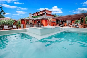 A swimming pool with a tiled deck, a covered seating area, and a building with a red facade and a thatched roof. - Azeda Boutique Hotel João Fernandes (Búzios)