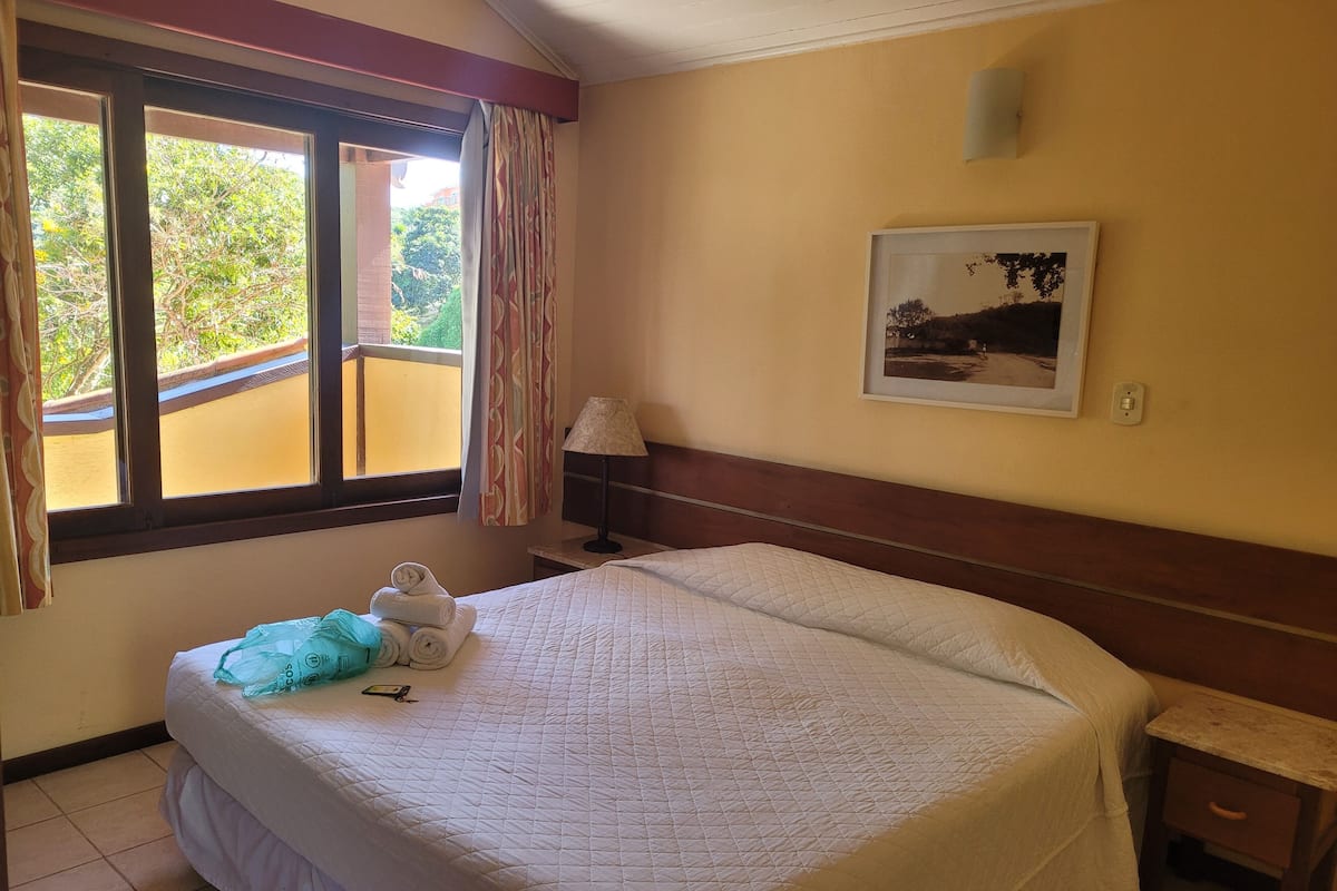 Una cama bien hecha con un edredón blanco acolchado, una lámpara de noche y una mesita de noche con lámpara. Hay una ventana con cortinas, un cuadro en la pared y vistas a zonas verdes.