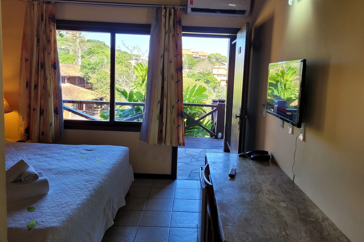 Habitación de hotel con cama, cortinas, televisor y balcón con vistas a zonas verdes y edificios.