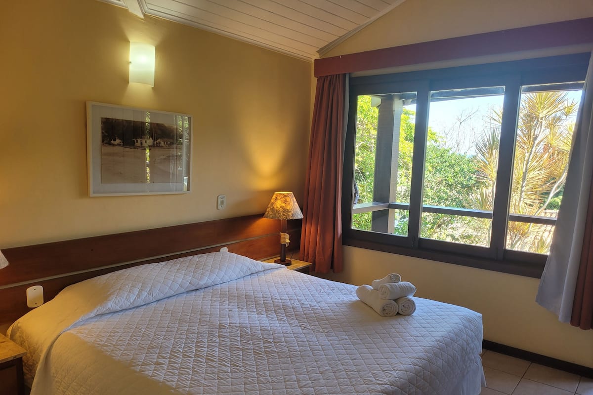 Habitación de hotel con una cama grande, una mesita de noche con lámpara, un cuadro enmarcado en la pared y una ventana con cortinas.