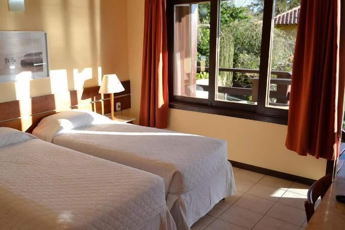 Habitación de hotel con dos camas, una ventana con cortinas, una lámpara y un cuadro colgado en la pared.